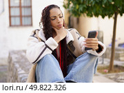 A young woman sits thoughtfully upon a sturdy stone wall, gazing intently at her smartphone while surrounded by a serene urban backdrop that enhances the beauty of her contemplative moment. A Thoughtful Young Woman is Engaged with Her Smartphone in a Bustling Urban Setting. Стоковое фото, агентство Ingram Publishing / Фотобанк Лори