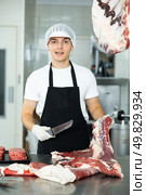 Positive young salesman cutting piece of meat in butcher shop. Стоковое фото, фотограф Яков Филимонов / Фотобанк Лори