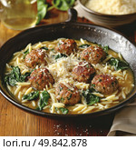 A mouth-watering dish served in a deep bowl: juicy meatballs with herbs, orzo (Italian pasta in the form of rice), fresh spinach leaves and broth, garnished with a generous portion of grated Parmesan cheese. The scene was filmed in a cozy rustic setting, on a wooden table. The dish looks rich and satisfying, perfect for autumn-winter menus, content about home cooking, recipes, food styling and gastronomic blogs. Стоковое AI-изображение, AI-художник Анна Кривицкая / Фотобанк Лори