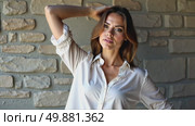 A woman in a white blouse standing confidently with her arm raised against a textured stone wall, soft lighting enhancing. Стоковое AI-видео, аниматор Sergey Grishin / Фотобанк Лори