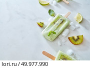Frozen green smoothies lolly with a slice of kiwi on a stick with mint leaves, ice and pieces of lime and kiwi on a gray marble table with space for text. Flat lay. Lime minty frozen smoothies on a stick with pieces of kiwi presented on a gray marble background with mint leaves and pieces of kiwi, lime and ice. Copy space. Flat lay. Стоковое фото, агентство Ingram Publishing / Фотобанк Лори