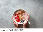 Female hands holding a plate with a fresh breakfast of yogurt, granola, ripe berries and almonds on a gray concrete background, top view.. Plate with a spoon and an apetitic yoghurt with berries and almonds women’s hands holding on a gray concrete background. Стоковое фото, агентство Ingram Publishing / Фотобанк Лори
