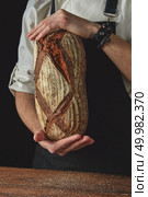 hands men keep the fresh bread of a dark apron and a wooden table on the black background. Hands of man holding oval bread. Стоковое фото, агентство Ingram Publishing / Фотобанк Лори