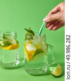 Citrus fruits slices of lemon and lime, ice, leaf of mint, water. Two glass jar on green background with plastic straws with famale hand. Cold natural lemonade handmade with bubbles of air.. A woman’s hand inserts plastic straws into a glass jar with lemonade from natural ingredients - water, mint, slices of lemon, lime. Natural cold lemonade handmade. Стоковое фото, агентство Ingram Publishing / Фотобанк Лори
