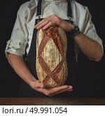 On a black background the hands man holding a fresh dark homemade bread. Hands of man holding oval bread. Стоковое фото, агентство Ingram Publishing / Фотобанк Лори