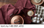 Copper saucepan, cooking utensils for cooking dishes with eggs in a cardboard box and flour in a bowl on a claret apron. Ingredients for pancakes or pie on old wooden background, flat lay. Kitchen utensils and ingredients for making a pie on an old wooden table with an apron. Стоковое фото, агентство Ingram Publishing / Фотобанк Лори