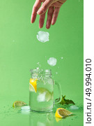Splashes and drops of a natural cocktail scatter on the surface of the table. A female hand throws ice cubes into a glass with a homemade natural cocktail on a green background.. A woman’s hand throws a piece of ice into a glass jar with a natural lemonade. Ice cubes fall into a glass jar with a splash and drops in different directions. Стоковое фото, агентство Ingram Publishing / Фотобанк Лори
