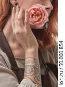Delicate pink rose in the mouth of a red-haired young girl with a tattoo. The concept of a holiday card. The red-haired girl with a tattoo is holding a pink rose in her mouth. Layout for postcard. Стоковое фото, агентство Ingram Publishing / Фотобанк Лори