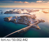 Aerial view of bridge over the sea and snowy mountains in Lofoten Islands, Norway. Henningsvaer at sunset in winter. landscape with blue water, sky with clouds, rocks, buildings, road. Top view. Стоковое фото, агентство Ingram Publishing / Фотобанк Лори