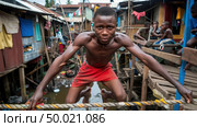 A young boy balances on a rope in front of a row of small shacks, showcasing his agility and skill in a challenging environment. Стоковое AI-изображение, AI-художник Данила Большаков / Фотобанк Лори