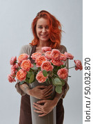 A smiling red-haired woman in a brown apron holds a vase with roses in a color of the year 2019 Living Coral Pantone on a gray background with space for text. The concept of a flower shop. Young woman with a big vase of roses flowers in a color of the year 2019 Living Coral Pantone on a gray background with copy space. Стоковое фото, агентство Ingram Publishing / Фотобанк Лори