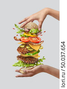 Big deliciouse homemade flying hamburger from organic vegetables and fresh natural beef cutlets in a woman’s hands on a white background. Place for text.. Girl’s hands hold big tasty homemade burger with flying natutal ingredients on a white background. Copy space. Стоковое фото, агентство Ingram Publishing / Фотобанк Лори
