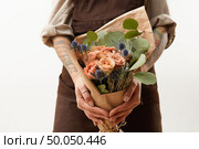 Young girl in a brown apron holds beautiful bunch with fresh natural flowers living coral colored and green leaves on a light background, copy space. Holiday congratulation card.. Close-up of greeting bouquet from fresh roses, eryngium and green leaves a in a female’s hands with tattoo on a gray background. Place for text. Gift for Mother’s Day. Стоковое фото, агентство Ingram Publishing / Фотобанк Лори