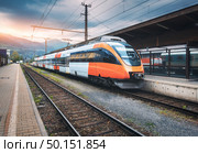 High speed train on the railway station in mountains at sunset in summer. Orange modern commuter train on the railway platform. Industrial landscape with railroad. Passenger transportation. Intercity. High speed train on the railway station in mountains at sunset. Стоковое фото, агентство Ingram Publishing / Фотобанк Лори