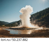 A stunning display of nature unfolds as a geyser erupts forcefully, sending a plume of steam high into the air against a backdrop of lush hills and a clear sky. Nature flourishes Generative AI. Стоковое AI-изображение, AI-художник Юлия Петровская / Фотобанк Лори
