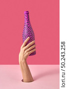 Painted hot pink wine bottle with black spots in a woman’s hand on a duotone pink background, copy space. Holiday concept.. Female’s hand with painted black spots champagne bottle. Стоковое фото, агентство Ingram Publishing / Фотобанк Лори