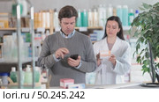 Attentive middle-aged man scrolling phone while looking for something and pharmacist consulting him in drugstore. Стоковое видео, видеограф Яков Филимонов / Фотобанк Лори