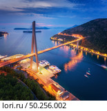 Aerial view of beautiful modern bridge at night. Dubrovnik, Croatia. Top view of road, boats, yachts and city lights at sunset. Summer landscape with harbor, mountain, highway, blue sea and cloudy sky. Стоковое фото, агентство Ingram Publishing / Фотобанк Лори