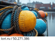A close-up of fishing floats and ropes against a seaport in overcast weather. In the center of the frame is a yellow float with raindrops, surrounded by blue and orange buoys and thick sea ropes. The background is slightly blurred, with the outlines of docks and buildings visible. The image conveys the atmosphere of maritime routine, fishing labor and coastal life. Ideal for the themes "maritime industry", "fishing", "port", "navigation", "tackle" and "atmospheric texture". Стоковое AI-изображение, AI-художник Анна Кривицкая / Фотобанк Лори
