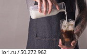 Barista with tattoo is pouring fresh milk from bottle into glass with fresh brewed coffee and ice cubes. 4K UHD video, 240fps, 2160p.. The man with tatoo makes refreshing ice coffee with milk. Стоковое фото, агентство Ingram Publishing / Фотобанк Лори