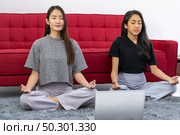 Two asian Woman do meditate from online meditation coaching to relax and reduce stress and strain for good mental health cause from city lockdown from coronavirus COVID-19 Pandemic. Стоковое фото, агентство Ingram Publishing / Фотобанк Лори