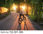 Woman riding a bicycle on the rural road at sunset in summer. Colorful landscape with sporty girl with backpack riding a mountain bike, road, green trees and gold sunlight. Sport and travel. Cycle. Стоковое фото, агентство Ingram Publishing / Фотобанк Лори