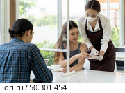 Asian waitress wear facemask and face shield using tablet tp show restaurant electronic menu and recommend to customer. New normal technology and lifestyle food and dining concept. Стоковое фото, агентство Ingram Publishing / Фотобанк Лори