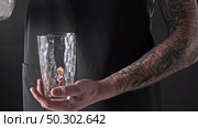 A barista man pours ice cubes into a glass with splashes of drops on a dark background. 4K UHD video, 240fps, 2160p.. A man with a tattoo is pouring ice cubes into a glass around a dark background. 4K UHD video, 2160p. Стоковое фото, агентство Ingram Publishing / Фотобанк Лори