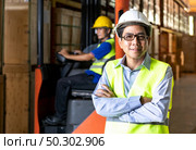 Portrait of Asian warehouse manager stand and arm crossed in distribution warehouse with warehouse worker operate forklift in background. Reopening business warehouse technology and logistic concept. Стоковое фото, агентство Ingram Publishing / Фотобанк Лори
