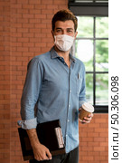 Portrait of middle east caucasian businessman with takeout coffee cup and clipboard in meeting room after lunch break. He wear face mask as new normal office preventing coronavirus COVID-19 spreading. Стоковое фото, агентство Ingram Publishing / Фотобанк Лори