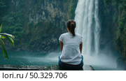 A woman is seated in front of a waterfall, watching the cascading water with awe and contemplation. Стоковое AI-изображение, AI-художник Данила Большаков / Фотобанк Лори