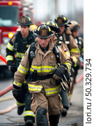 Firefighters in full gear walking down a city street, likely on their way to respond to an emergency. They are wearing helmets, jackets, and carrying various equipment. Стоковое AI-изображение, AI-художник Данила Большаков / Фотобанк Лори