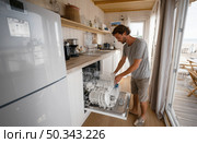 Man unloading dishwasher in bright coastal kitchen, casual home setting, daylight, modern interior, wooden floor, and beach view through glass doors. Стоковое AI-изображение, AI-художник Катерина Бондарец / Фотобанк Лори