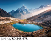 Snowy mountain with illuminated peaks and small lake with blue water at sunrise in Nepal. Beautiful landscape with lake, high rocks in snow, golden sun rays, hills at dawn. Himalayan mountains. Nature. Стоковое фото, агентство Ingram Publishing / Фотобанк Лори