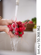 Close up view of a woman washes a bunch of red radishes under the pressure of flowing water in the white kitchen, a basket with tomato, broccoli and onions in the background, grapes. Стоковое фото, фотограф Vladimir Drozdin / Фотобанк Лори