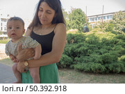 Молодая женщина держит на руках четырёхмесячную девочку в полосатой футболке на фоне густой зелёной изгороди в солнечном парке. Стоковое фото, фотограф Владислав Войтов / Фотобанк Лори