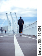 05/02/2025 Russia.Moscow.Person in dark clothing rides an electric scooter on a bridge pathway. Background includes blurred figures, tall structures, and a cloudy sky. Редакционное фото, фотограф Алина Лебедь / Фотобанк Лори