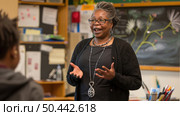 Mature African american woman is engaged in conversation with a child in a classroom setting, surrounded by desks and educational materials. Стоковое AI-изображение, AI-художник Данила Большаков / Фотобанк Лори