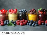 A bright and appetizing image of an assortment of berry smoothies and mousses served in glass jars. Each jar contains a multi-colored puree or drink, decorated with fresh berries: blueberries, raspberries, blackberries, red currants. In front of the jars, in the background, there are berries and mint leaves. The background is dark, which emphasizes the rich colors of the ingredients. The photo was taken in a studio with natural light, ideal for nutrition, healthy lifestyle, veganism, detox diets and food photography. Стоковое AI-изображение, AI-художник Анна Кривицкая / Фотобанк Лори