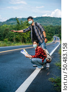 Female tourists sit and look at the map, male tourists pretending to hitchhike. Both wear masks and are on the side of the road. Стоковое фото, агентство Ingram Publishing / Фотобанк Лори