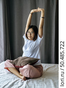 A woman in a white shirt sitting on the bed and raising both arms. Стоковое фото, агентство Ingram Publishing / Фотобанк Лори