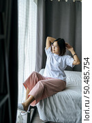 A woman in a white shirt sitting on the bed and raising both arms. Стоковое фото, агентство Ingram Publishing / Фотобанк Лори