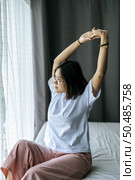 A woman in a white shirt sitting on the bed and raising both arms. Стоковое фото, агентство Ingram Publishing / Фотобанк Лори
