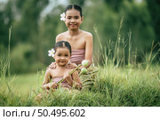 Close up, Portrait of Cute sister and young sister in Thai traditional dress and put white flower on her ear sitting in meadow,  smile and looking at camera, sibling love concept, copy space. Стоковое фото, агентство Ingram Publishing / Фотобанк Лори