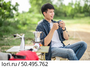 Young traveler man hold coffee cup and sitting at front of the tent in forest while camping trip on summer vacation with coffee set and making fresh coffee grinder. Стоковое фото, агентство Ingram Publishing / Фотобанк Лори