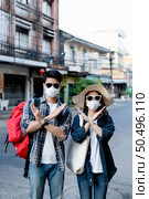 Young backpacker couple in face mask and sunglasses make a prohibition cross mark with both hands express negative attitude coronavirus don’t close contact while travel in city street On City Street. Стоковое фото, агентство Ingram Publishing / Фотобанк Лори