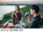 Selective focus,  Pretty girl smiling,  Asian young couple sitting drinking coffee to happily near lake and front of tent on camping trip. Lifestyle and camping concept. Стоковое фото, агентство Ingram Publishing / Фотобанк Лори