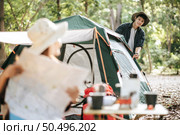 Selective focus, Pretty woman sitting on chair at front of camping tent and checking direction on paper map, Handsome boyfriend pitching a tent behind her, They happy to camping in forest on vacation. Стоковое фото, агентство Ingram Publishing / Фотобанк Лори