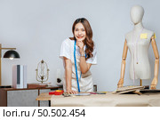 Portrait of young pretty female fashion designer stylish standing and hand on shin with smile feeling proud her business during working at fashion studio, full of tailoring tools and equipment. Стоковое фото, агентство Ingram Publishing / Фотобанк Лори