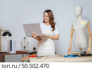 Beautiful young Asian fashion designer or tailor with working with laptop computer to search idea or online orders,  full of tailoring tools and equipment on the desk in the studio. Стоковое фото, агентство Ingram Publishing / Фотобанк Лори